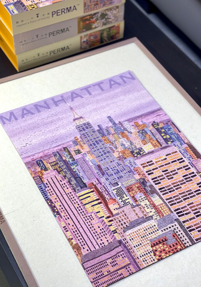 Finished Fantasy Manhattan puzzle laid flat on a table with stacked PERMA puzzle boxes in the background.