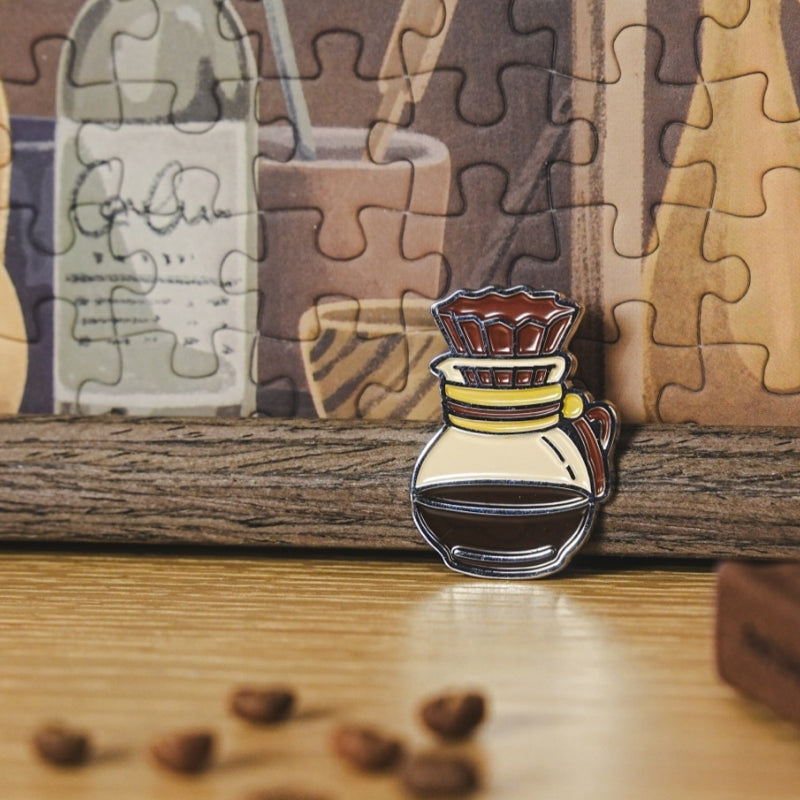 Close-up of enamel pin shaped like a coffee pot in front of puzzle.