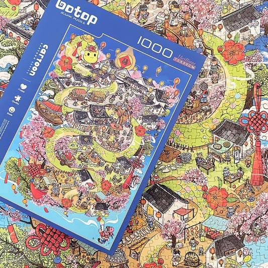 Assembled Draw Snake puzzle next to its box, featuring colourful village streets, blossom trees and market stalls