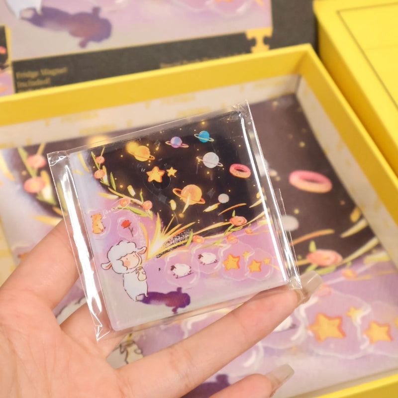 Close-up of a hand holding the fridge magnet featuring the ‘Unlock the Universe’ lamb releasing glowing stars and planets.