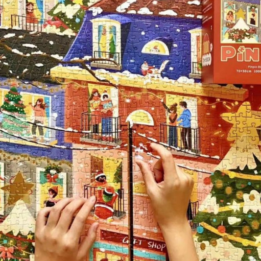Hands assembling Pinpin Blessed Night 1000 piece jigsaw puzzle with festive holiday scene and decorated houses.